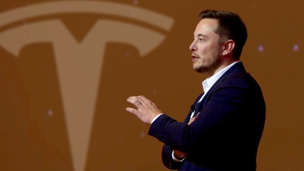 Tesla CEO Elon Musk giving a presentation (right), large Tesla logo in the background (left).