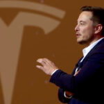 Tesla CEO Elon Musk giving a presentation (right), large Tesla logo in the background (left).
