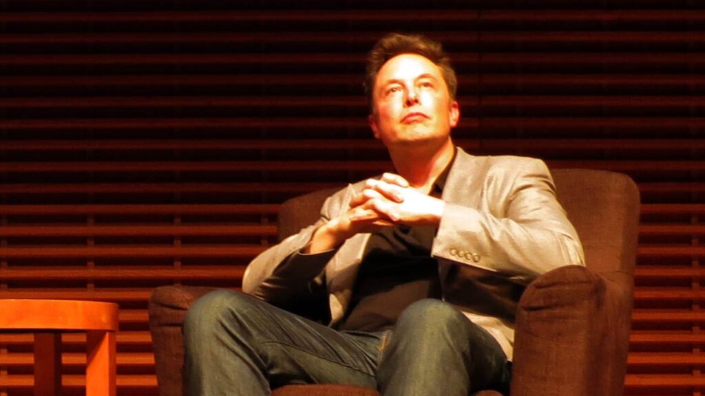 Elon Musk sitting as a guest at a conference.