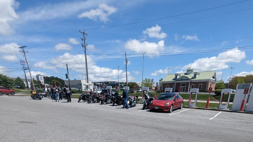 A gang of bikers swarms the Tesla Supercharger station as the lone Tesla Model 3 owner returns to his car after a charging break.