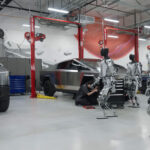 Multiple Tesla Optimus humanoid robots walk inside a Tesla factory workshop (Cybertruck prototypes can be seen in the background).