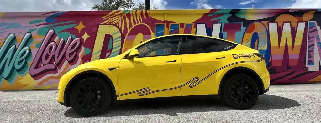 DASH ridesharing Tesla Model Y wrapped in yellow. The wall in background painted in graffiti saying "We Love Downtown".