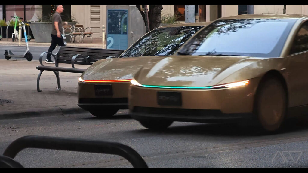 Two Tesla Cybercab robotaxis on the streets of Austin, Texas, as one shows an orange front light bar and the other has an aqua blue color.
