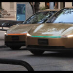 Two Tesla Cybercab robotaxis on the streets of Austin, Texas, as one shows an orange front light bar and the other has an aqua blue color.