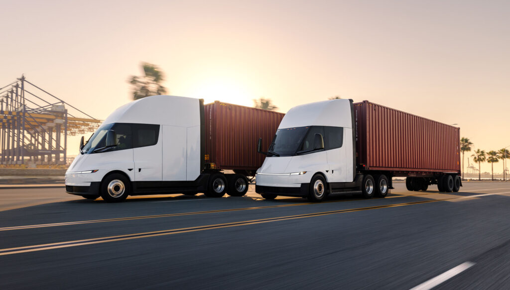 Two Tesla Semi trucks hauling shipping containers. The one in front is the Standard Range (325 miles), and the second one behind it is the Long Range (500-mile) variant (extended cabin and roof fairing).